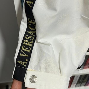 May include: A white jacket sleeve featuring a black vertical stripe with the gold text A. VERSACE. The fabric has a subtle tonal embossed pattern and a single silver-toned button on the cuff. The design includes a classic collar-like cuff detail.