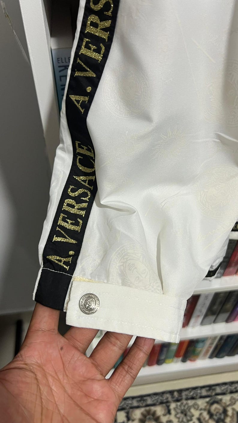 May include: A white jacket sleeve featuring a black vertical stripe with the gold text A. VERSACE. The fabric has a subtle tonal embossed pattern and a single silver-toned button on the cuff. The design includes a classic collar-like cuff detail.