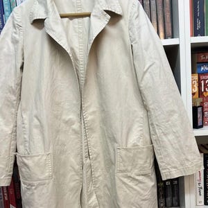 Puede incluir: Una chaqueta de color beige claro con mangas largas, cuello cl&aacute;sico, cierre frontal de botones y dos grandes bolsillos cuadrados de parche situados en los paneles inferiores delanteros. La prenda se muestra colgada en una percha de madera.