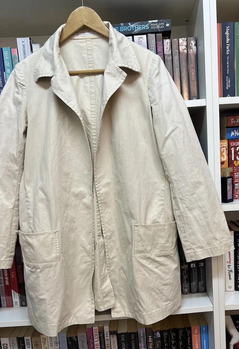 Puede incluir: Una chaqueta de color beige claro con mangas largas, cuello cl&aacute;sico, cierre frontal de botones y dos grandes bolsillos cuadrados de parche situados en los paneles inferiores delanteros. La prenda se muestra colgada en una percha de madera.
