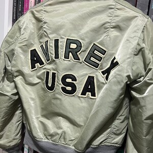 May include: A sage green bomber jacket featuring a gray ribbed collar and cuffs. The back displays large, black and white embroidered lettering that reads AVIREX USA in a curved arrangement, with the word USA positioned below the word AVIREX.