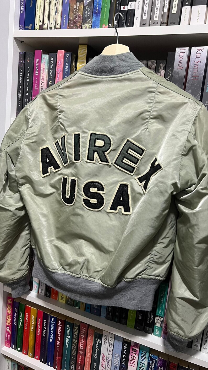 May include: A sage green bomber jacket featuring a gray ribbed collar and cuffs. The back displays large, black and white embroidered lettering that reads AVIREX USA in a curved arrangement, with the word USA positioned below the word AVIREX.