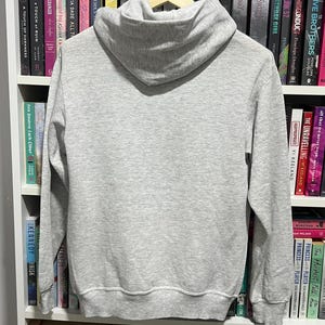 May include: A light grey long-sleeve pullover hoodie made of a soft fabric, featuring a hood, ribbed cuffs at the wrists, and a ribbed hem at the bottom. The hoodie is displayed hanging on a wooden hanger, showing the back side of the garment.