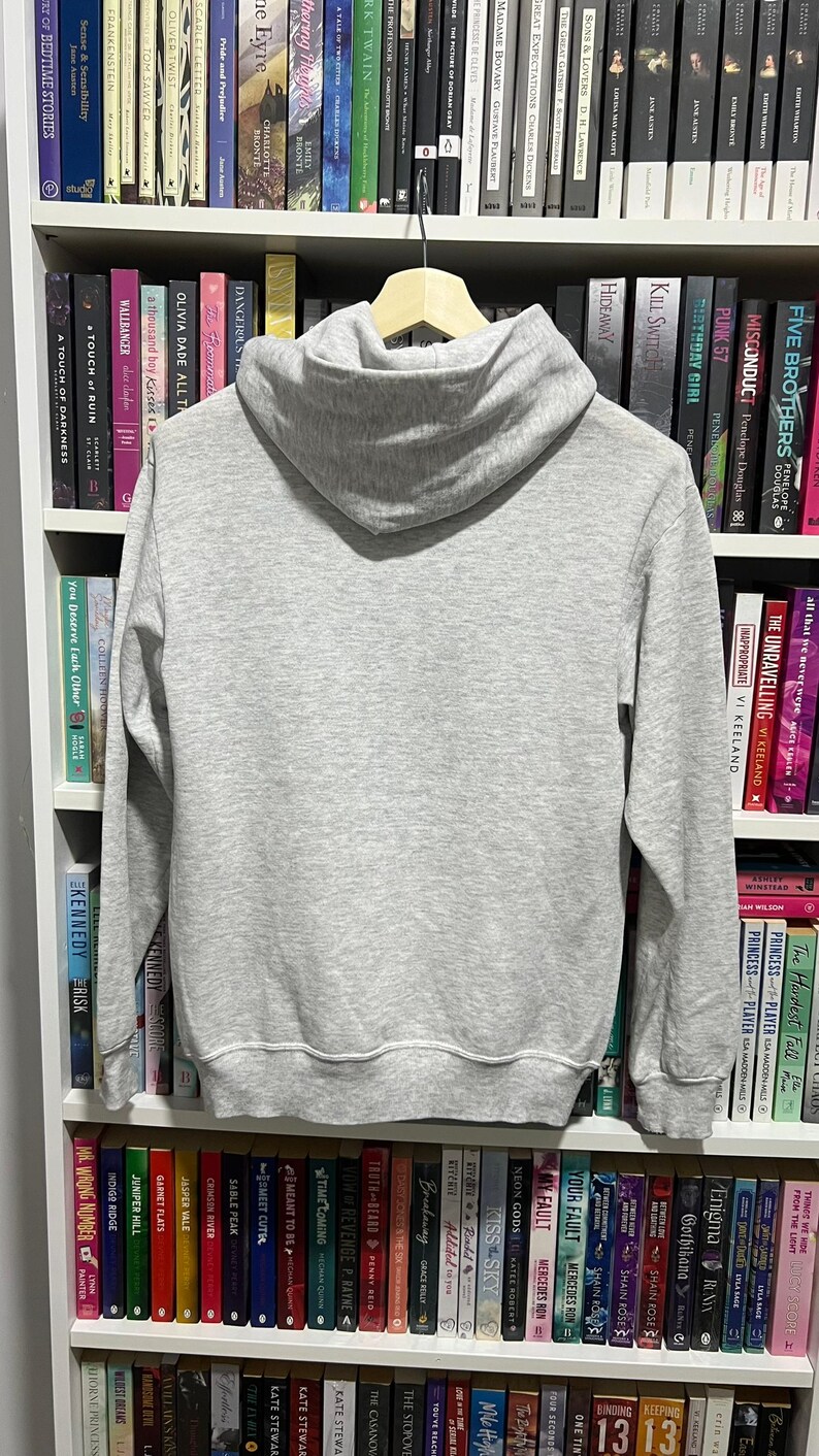 May include: A light grey long-sleeve pullover hoodie made of a soft fabric, featuring a hood, ribbed cuffs at the wrists, and a ribbed hem at the bottom. The hoodie is displayed hanging on a wooden hanger, showing the back side of the garment.