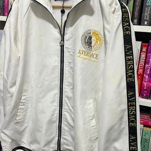May include: A white zip-up jacket featuring a black collar and black side stripes with the repeated text A. VERSACE. The chest displays a circular gold and silver emblem with the text AVERSACE. It includes a full-length black zipper and white drawstrings.