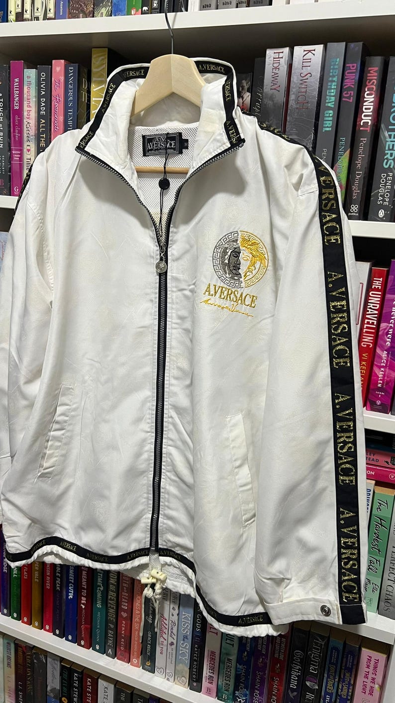 May include: A white zip-up jacket featuring a black collar and black side stripes with the repeated text A. VERSACE. The chest displays a circular gold and silver emblem with the text AVERSACE. It includes a full-length black zipper and white drawstrings.