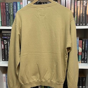 May include: A tan-colored sweatshirt with a crew neck and long sleeves hangs on a wooden hanger. The back of the sweatshirt is visible, showing a simple, minimalist design. The garment is made of a soft, comfortable material.