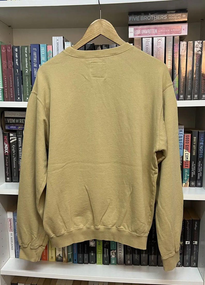 May include: A tan-colored sweatshirt with a crew neck and long sleeves hangs on a wooden hanger. The back of the sweatshirt is visible, showing a simple, minimalist design. The garment is made of a soft, comfortable material.