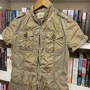 Puede incluir: Una chaqueta utilitaria de color caqui con mangas cortas, cremallera frontal completa, cuello alzado y dos bolsillos abotonados en el pecho. La prenda incluye charreteras en los hombros, cintura con cord&oacute;n ajustable y dos bolsillos inferiores.