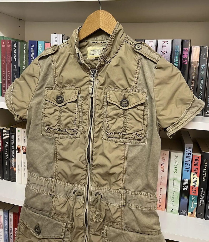 Puede incluir: Una chaqueta utilitaria de color caqui con mangas cortas, cremallera frontal completa, cuello alzado y dos bolsillos abotonados en el pecho. La prenda incluye charreteras en los hombros, cintura con cord&oacute;n ajustable y dos bolsillos inferiores.