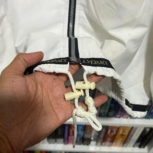 May include: A white jacket featuring a black centre zip and a black hem band with the text VERSACE A.VERSACE printed in gold. The jacket includes a white drawstring with two cream-coloured plastic cord locks at the bottom hem for adjustment.