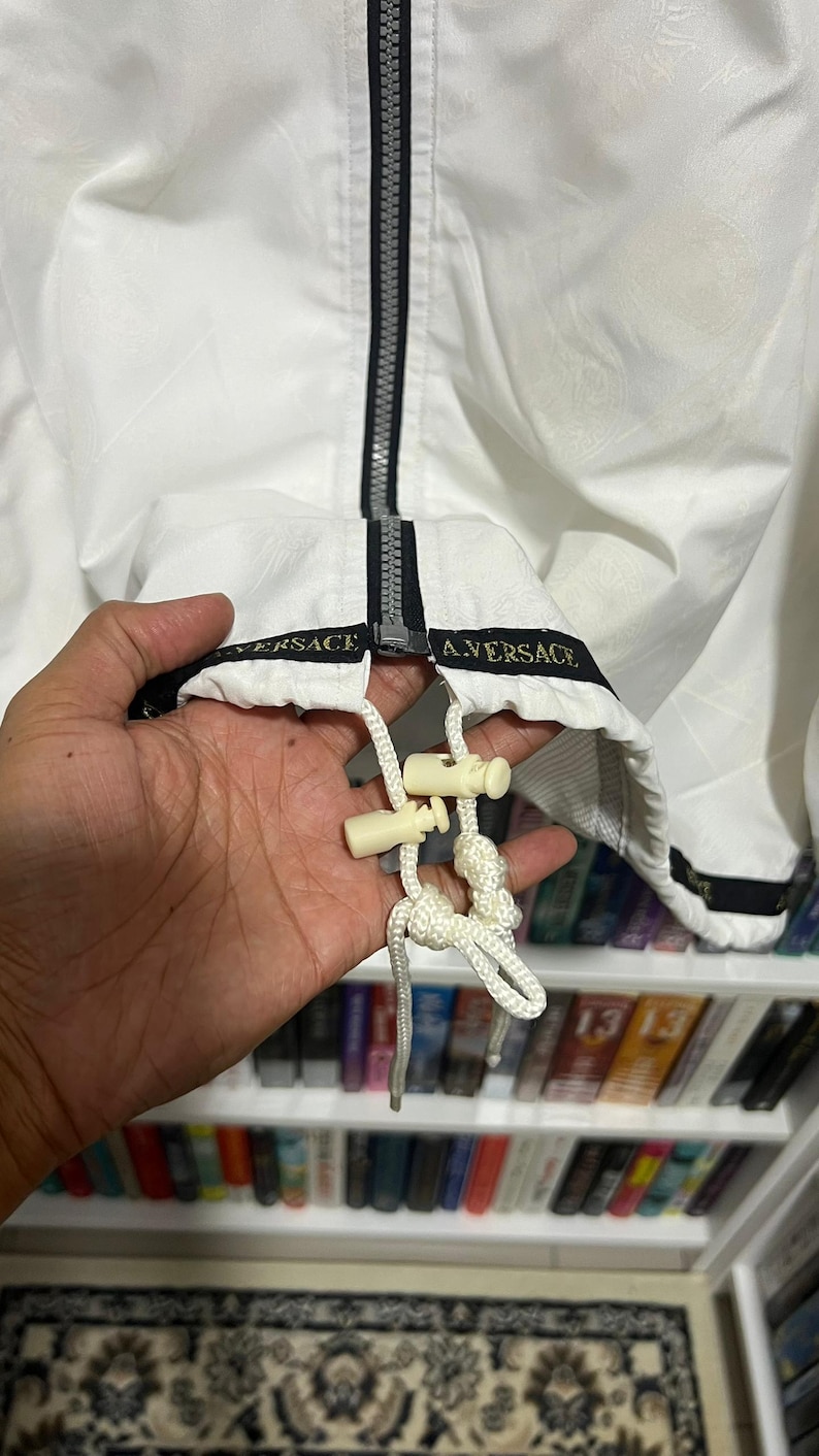 May include: A white jacket featuring a black centre zip and a black hem band with the text VERSACE A.VERSACE printed in gold. The jacket includes a white drawstring with two cream-coloured plastic cord locks at the bottom hem for adjustment.