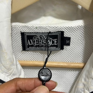 May include: A white jacket with a black zip and a black interior label that reads A.V. VERSACE NEW YORK A.V. VERSACE DIFFUSION with a size M tag. A black circular tag with the A.V. VERSACE logo hangs from the label by a thin black string.