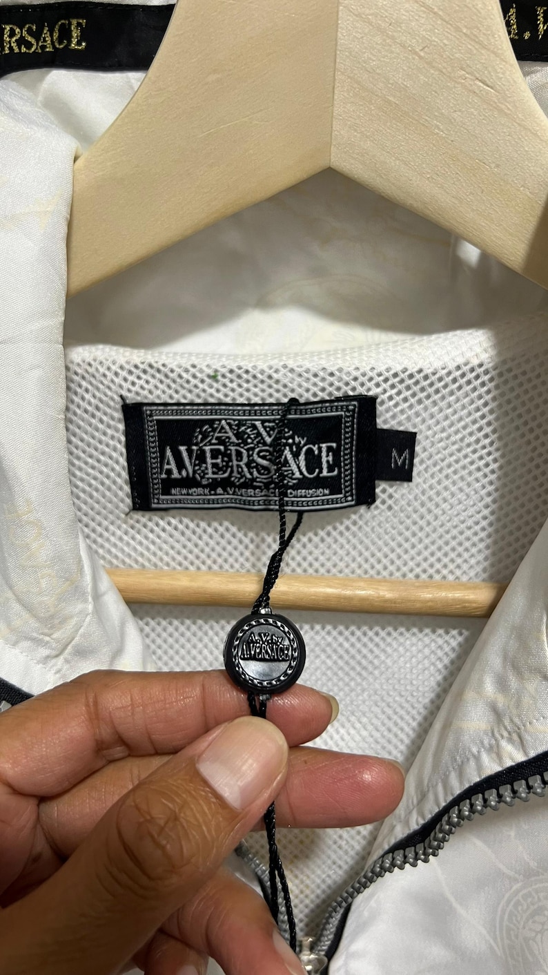 May include: A white jacket with a black zip and a black interior label that reads A.V. VERSACE NEW YORK A.V. VERSACE DIFFUSION with a size M tag. A black circular tag with the A.V. VERSACE logo hangs from the label by a thin black string.