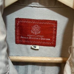 May include: Close-up of a tan shirt with a red label that reads "REGULATION AVIREX OFFICER'S UNIFORM." The shirt is hanging on a wooden hanger. A small white tag with the letter "M" is visible.