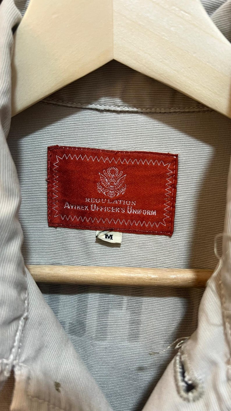 May include: Close-up of a tan shirt with a red label that reads "REGULATION AVIREX OFFICER'S UNIFORM." The shirt is hanging on a wooden hanger. A small white tag with the letter "M" is visible.