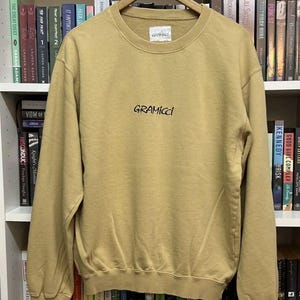 May include: A tan Gramicci sweatshirt with a crew neck and long sleeves. The brand name is printed in black script across the chest. The sweatshirt is hanging on a wooden hanger against a backdrop of bookshelves.