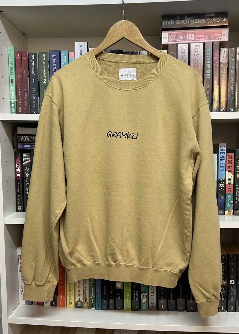May include: A tan Gramicci sweatshirt with a crew neck and long sleeves. The brand name is printed in black script across the chest. The sweatshirt is hanging on a wooden hanger against a backdrop of bookshelves.
