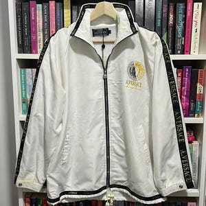 May include: A white zip-up track jacket featuring a black collar and hem with the text A.VERSACE repeated. Black stripes run down the sleeves with the same text. A circular logo with a split black and gold design sits on the chest with A.VERSACE.