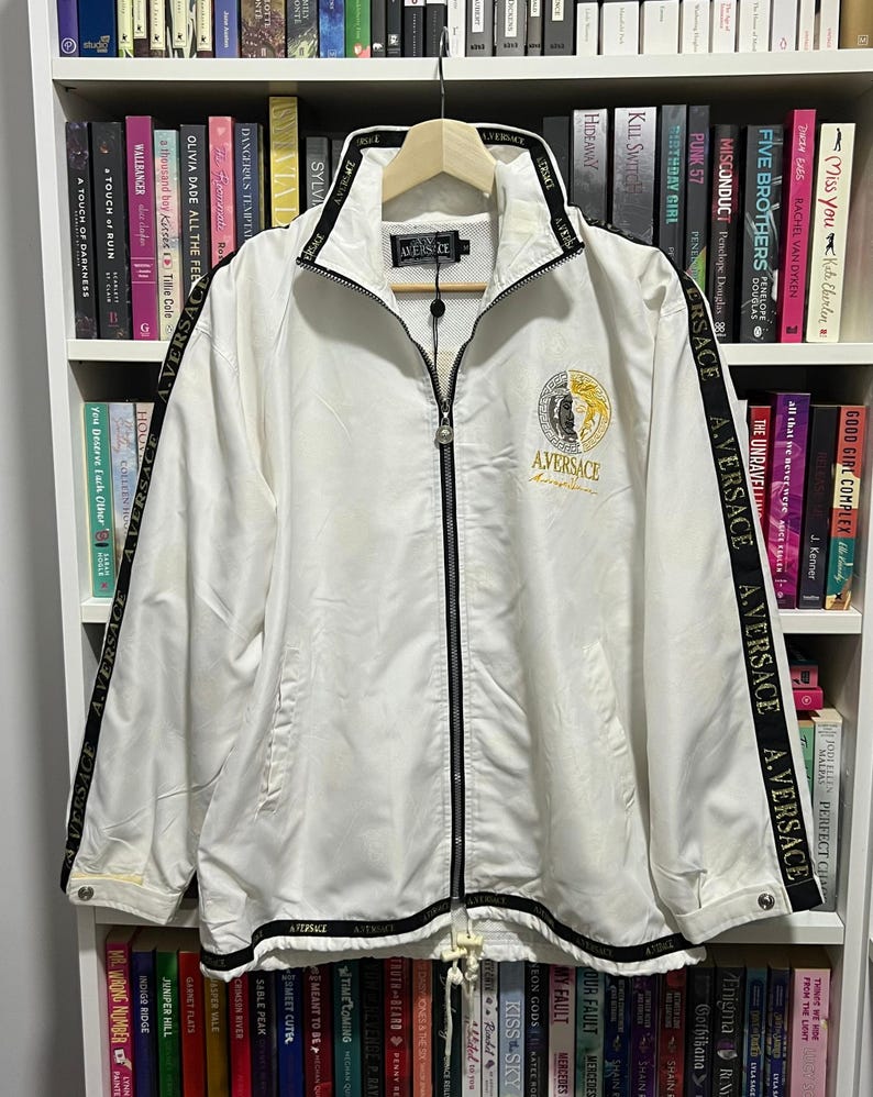 May include: A white zip-up track jacket featuring a black collar and hem with the text A.VERSACE repeated. Black stripes run down the sleeves with the same text. A circular logo with a split black and gold design sits on the chest with A.VERSACE.
