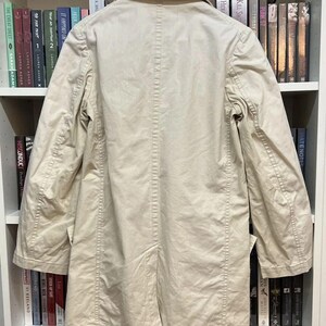 Puede incluir: Una gabardina de color beige con cuello cl&aacute;sico, mangas largas y una costura central en la espalda. La prenda se muestra en una percha de madera, destacando un dise&ntilde;o estructurado con costuras visibles a lo largo de los hombros y mangas.