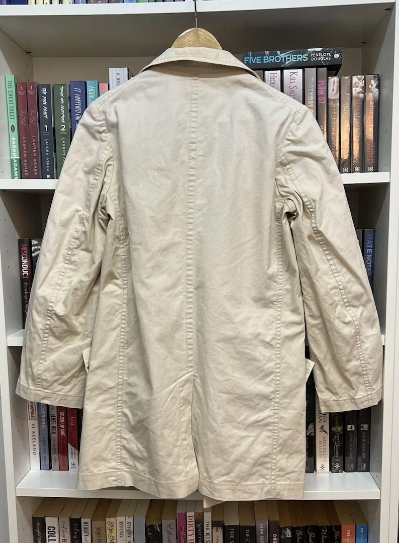 Puede incluir: Una gabardina de color beige con cuello cl&aacute;sico, mangas largas y una costura central en la espalda. La prenda se muestra en una percha de madera, destacando un dise&ntilde;o estructurado con costuras visibles a lo largo de los hombros y mangas.