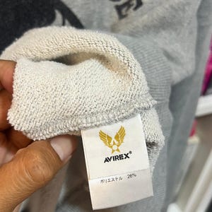 May include: Close-up of a gray sweatshirt with the word "FORCE" printed in black. The tag shows the brand "AVIREX&reg;" with a gold emblem and the text "ポリエステル 26%."