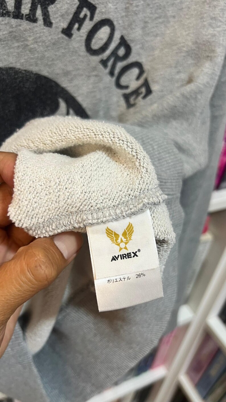 May include: Close-up of a gray sweatshirt with the word "FORCE" printed in black. The tag shows the brand "AVIREX&reg;" with a gold emblem and the text "ポリエステル 26%."