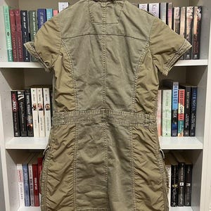 Puede incluir: Un vestido de estilo utilitario en color verde oliva con mangas cortas, cuello, cierre frontal de botones y cintura entallada. La prenda incluye m&uacute;ltiples bolsillos, costuras visibles y un dobladillo recto con cordones ajustables en la parte inferior.