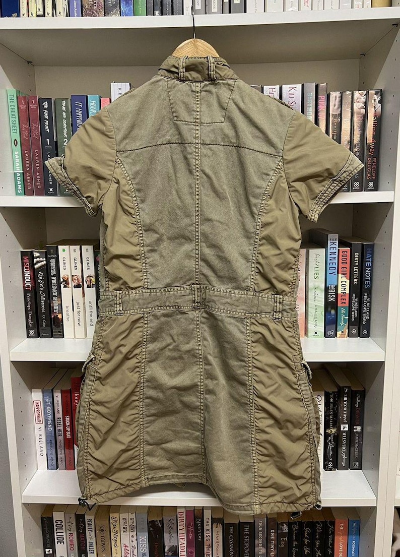 Puede incluir: Un vestido de estilo utilitario en color verde oliva con mangas cortas, cuello, cierre frontal de botones y cintura entallada. La prenda incluye m&uacute;ltiples bolsillos, costuras visibles y un dobladillo recto con cordones ajustables en la parte inferior.