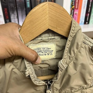 Puede incluir: Una chaqueta de color tostado con cuello alzado y cremallera frontal. Una etiqueta rectangular en el interior del cuello dice: Certificate of Authenticity EQUIPMENT-TYPE-AVIREX STOCK NO. 28-A6755 GARMENT SPECIFICATION SPEED LOW LEVEL FLIGHT AVIREX LTD EST 1975.