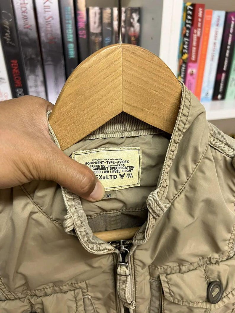 Puede incluir: Una chaqueta de color tostado con cuello alzado y cremallera frontal. Una etiqueta rectangular en el interior del cuello dice: Certificate of Authenticity EQUIPMENT-TYPE-AVIREX STOCK NO. 28-A6755 GARMENT SPECIFICATION SPEED LOW LEVEL FLIGHT AVIREX LTD EST 1975.