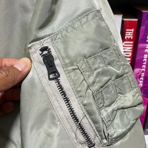 May include: A sage green bomber jacket sleeve featuring a utility pocket on the upper arm. The pocket includes a vertical black zipper with a branded metal pull and a rectangular flap-covered compartment with stitched detailing on the outer side.