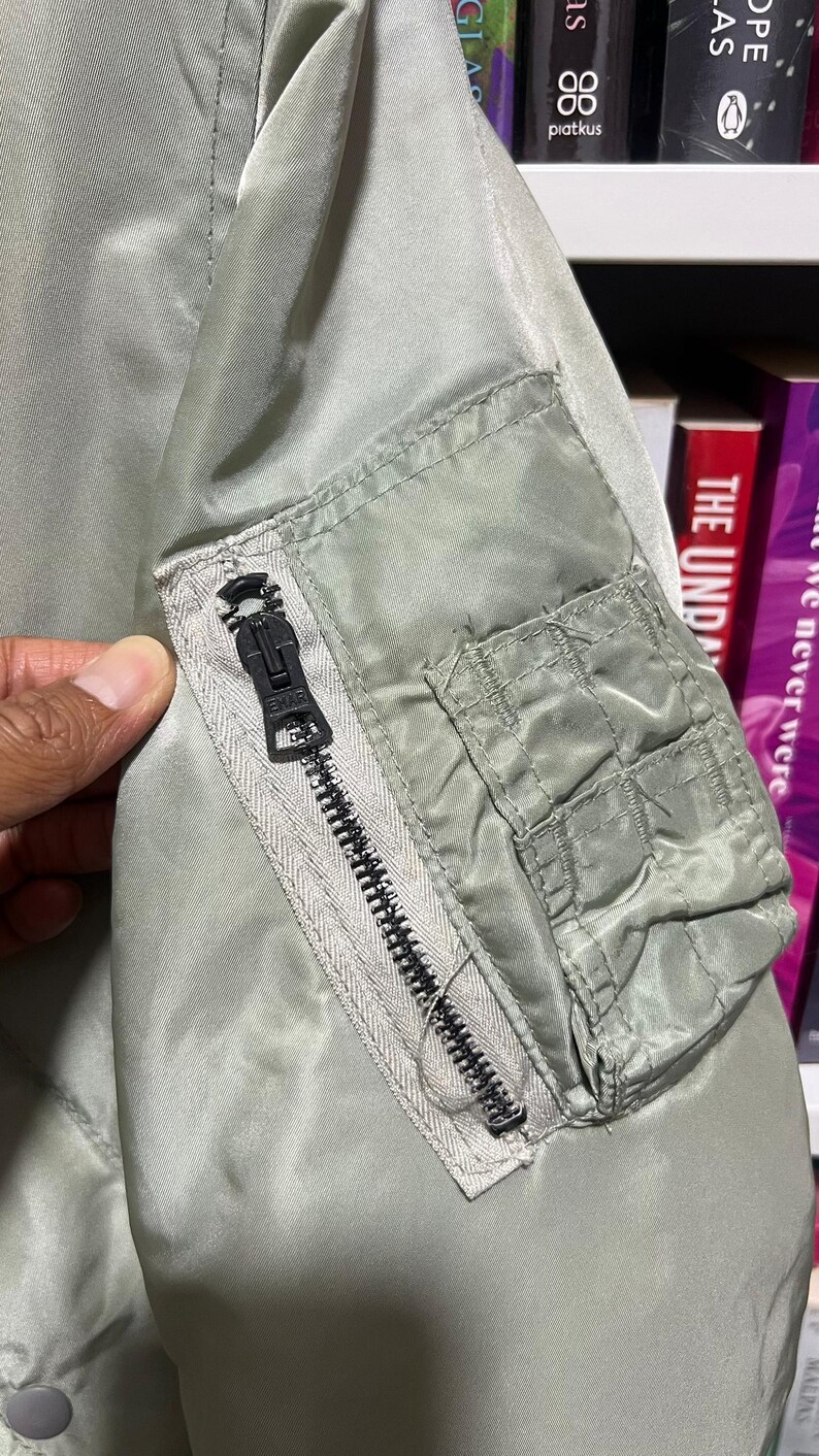 May include: A sage green bomber jacket sleeve featuring a utility pocket on the upper arm. The pocket includes a vertical black zipper with a branded metal pull and a rectangular flap-covered compartment with stitched detailing on the outer side.