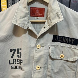 May include: A light tan military-style shirt with a collared neckline and button-up front. Features a "U.S. NAVY" patch, a "75 LRSP SQDRN" graphic, and an "AVIREX" patch. The shirt is hanging on a wooden hanger.