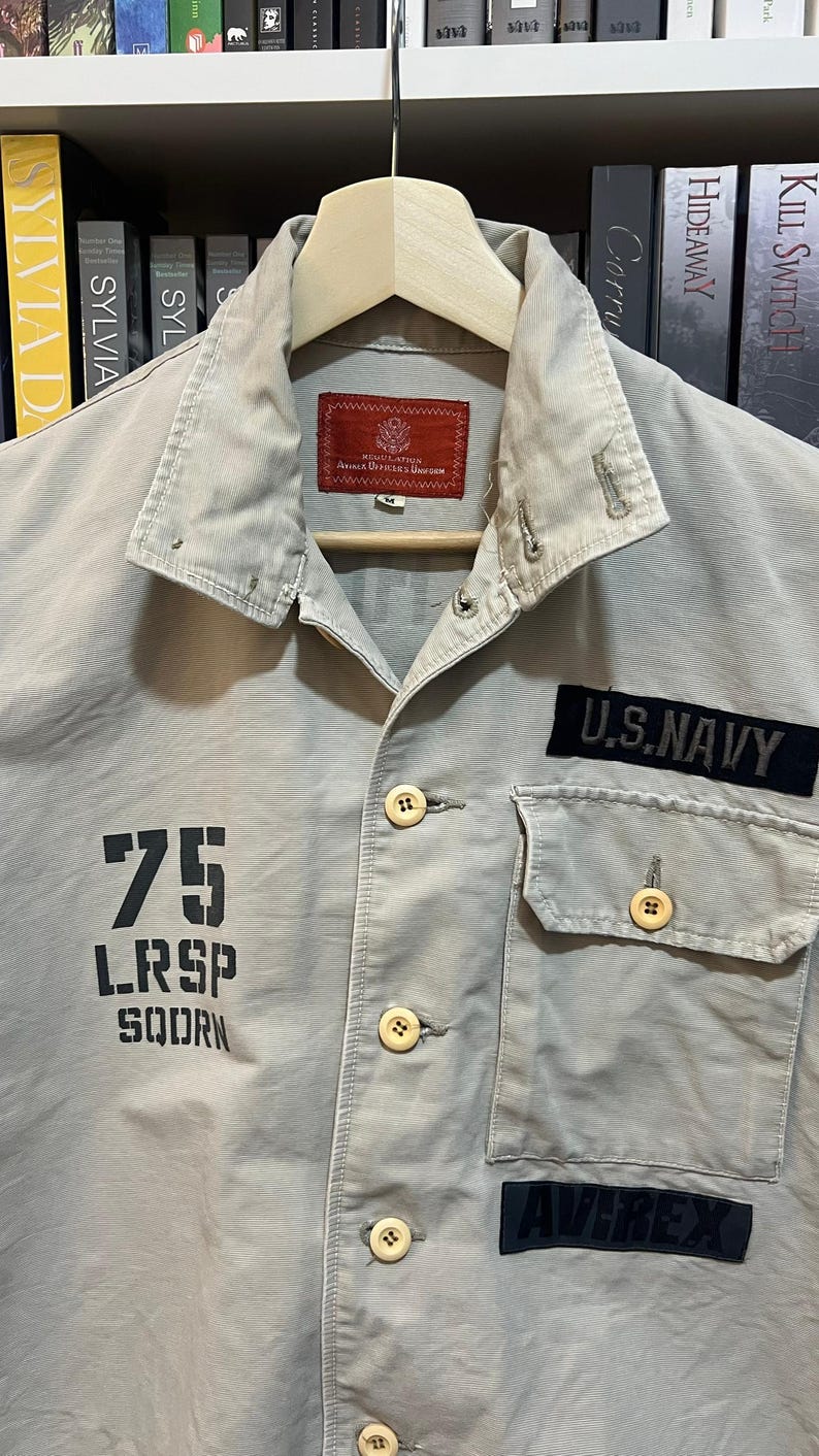 May include: A light tan military-style shirt with a collared neckline and button-up front. Features a "U.S. NAVY" patch, a "75 LRSP SQDRN" graphic, and an "AVIREX" patch. The shirt is hanging on a wooden hanger.