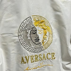 May include: A white jacket featuring a large circular embroidered emblem on the back. The emblem is split between silver and gold, depicting a face surrounded by a Greek key pattern. Below the emblem, the text AVERSACE is embroidered in gold.