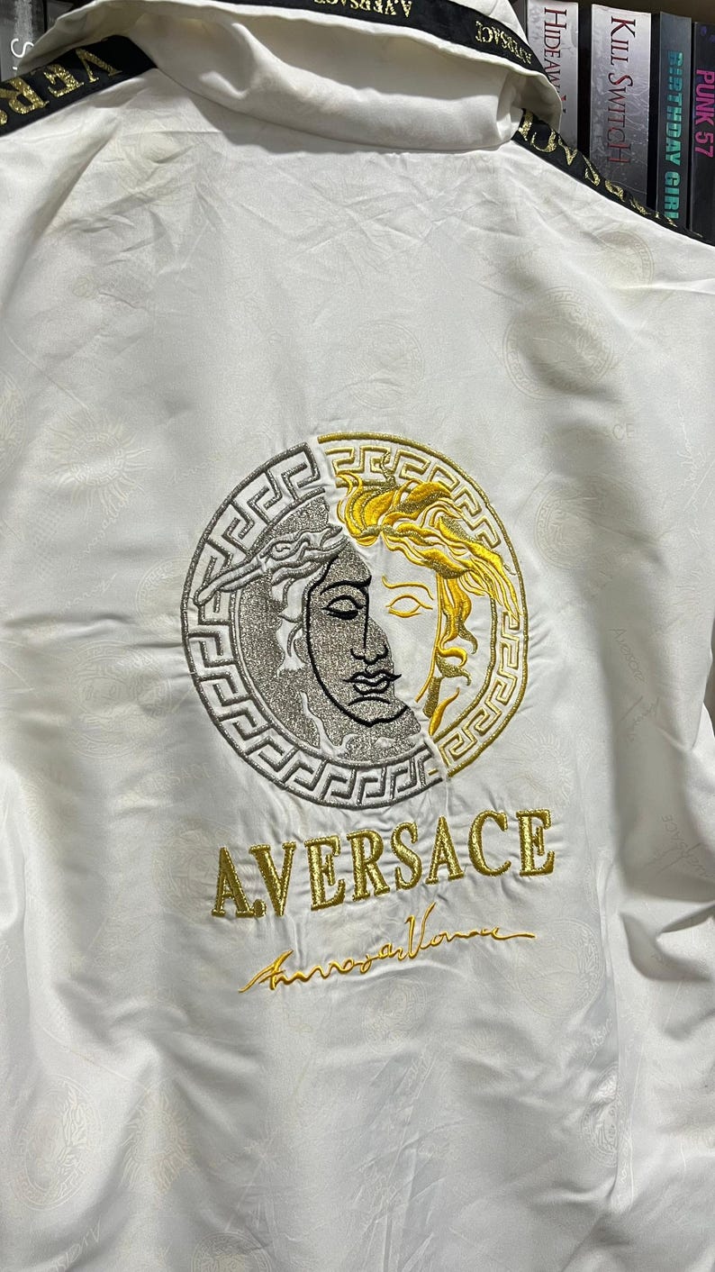 May include: A white jacket featuring a large circular embroidered emblem on the back. The emblem is split between silver and gold, depicting a face surrounded by a Greek key pattern. Below the emblem, the text AVERSACE is embroidered in gold.