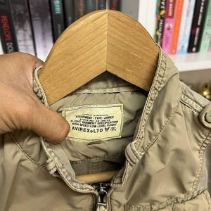 Puede incluir: Una chaqueta color canela con cremallera, cuello alto y bolsillos delanteros. Una etiqueta rectangular en el interior del cuello dice: Certificate of Authenticity EQUIPMENT-TYPE-AVIREX STOCK NO. 20-A6755 FLIGHT GARMENT SPECIFICATION HIGH SPEED LOW LEVEL FLIGHT AVIREX LTD EST 1975.