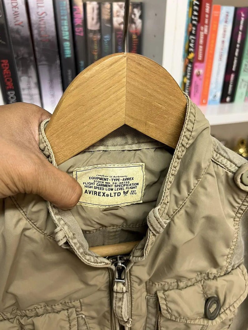 Puede incluir: Una chaqueta color canela con cremallera, cuello alto y bolsillos delanteros. Una etiqueta rectangular en el interior del cuello dice: Certificate of Authenticity EQUIPMENT-TYPE-AVIREX STOCK NO. 20-A6755 FLIGHT GARMENT SPECIFICATION HIGH SPEED LOW LEVEL FLIGHT AVIREX LTD EST 1975.