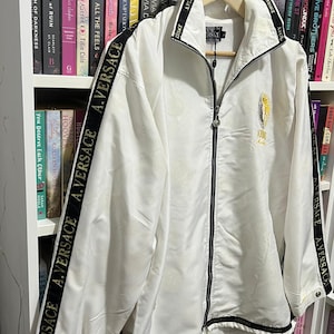 May include: A white zip-up jacket featuring a high collar and long sleeves with black stripes running down the arms. The black stripes are printed with the white text A.VERSACE. A gold circular logo is embroidered on the left chest area.