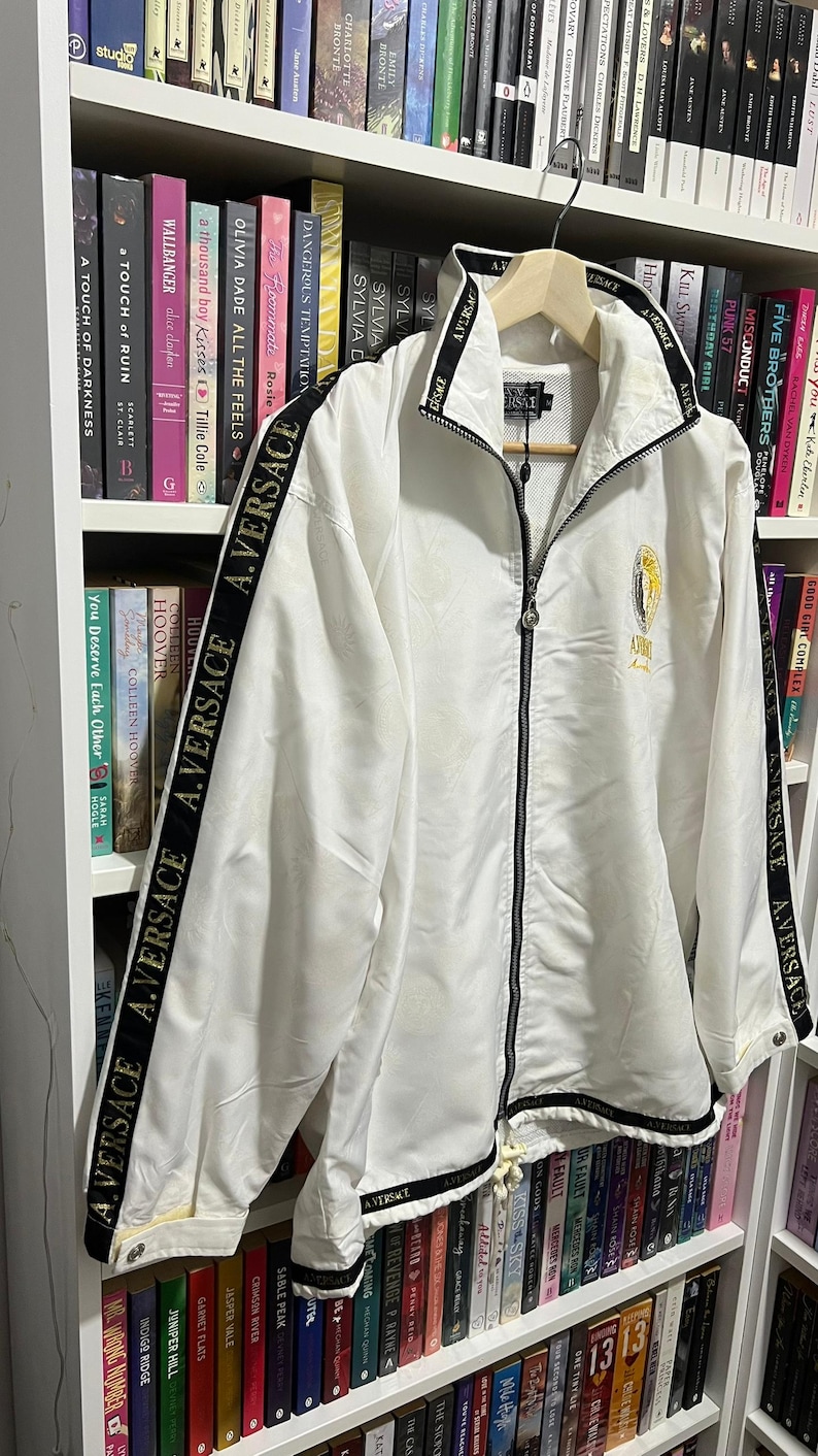 May include: A white zip-up jacket featuring a high collar and long sleeves with black stripes running down the arms. The black stripes are printed with the white text A.VERSACE. A gold circular logo is embroidered on the left chest area.