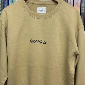 May include: A tan Gramicci sweatshirt with a crew neck and long sleeves. The brand name is printed in black script across the chest. The sweatshirt is hanging on a wooden hanger.