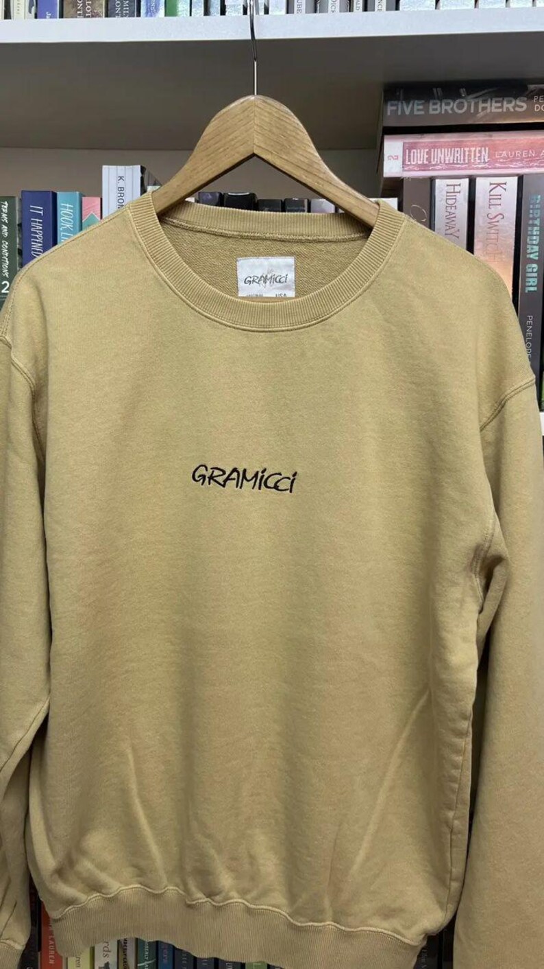 May include: A tan Gramicci sweatshirt with a crew neck and long sleeves. The brand name is printed in black script across the chest. The sweatshirt is hanging on a wooden hanger.