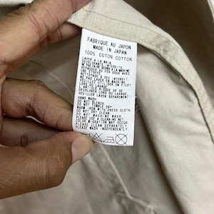 Puede incluir: Una etiqueta de cuidado de tela blanca cosida en una prenda beige. La etiqueta presenta texto negro que indica "MADE IN JAPAN" y "100% COTTON", junto con instrucciones detalladas de lavado a mano en franc&eacute;s e ingl&eacute;s, adem&aacute;s de cuatro s&iacute;mbolos de cuidado.