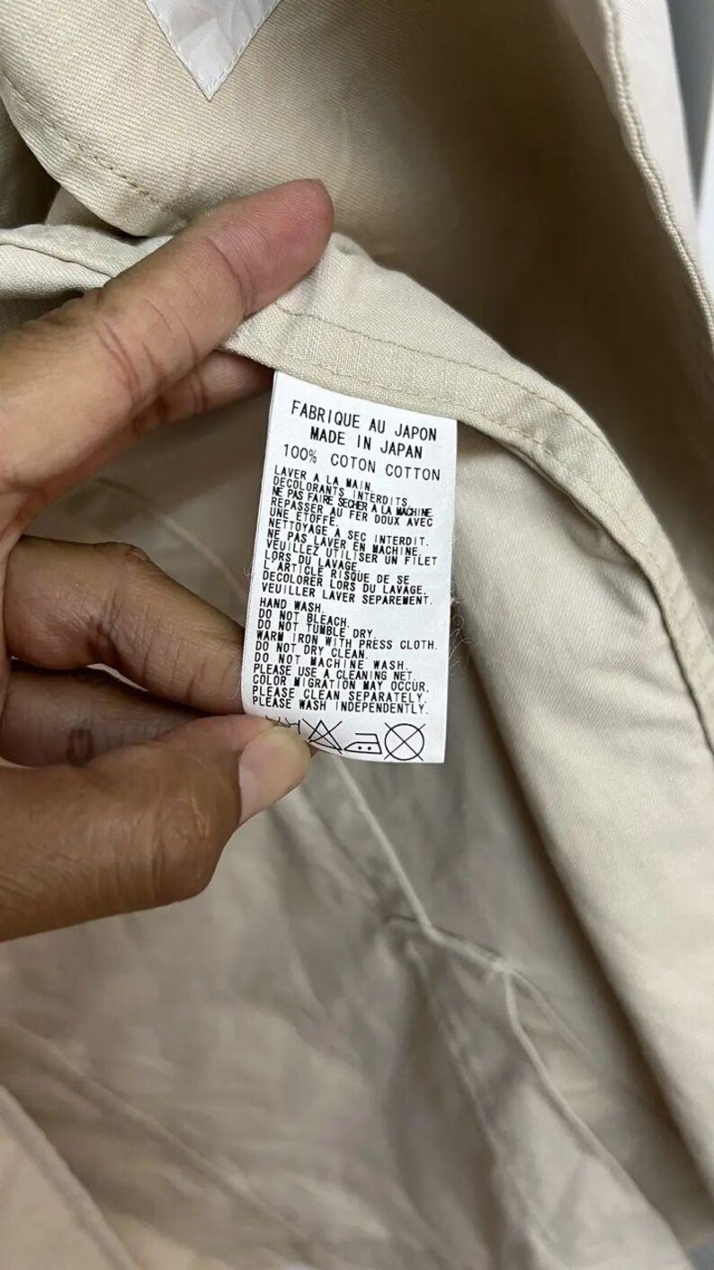 Puede incluir: Una etiqueta de cuidado de tela blanca cosida en una prenda beige. La etiqueta presenta texto negro que indica "MADE IN JAPAN" y "100% COTTON", junto con instrucciones detalladas de lavado a mano en franc&eacute;s e ingl&eacute;s, adem&aacute;s de cuatro s&iacute;mbolos de cuidado.