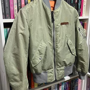 May include: A sage green bomber jacket featuring a ribbed gray collar, cuffs, and hem. The jacket has a full-length front zipper, two side pockets with snap closures, a small zippered utility pocket on the left sleeve, and a rectangular patch.