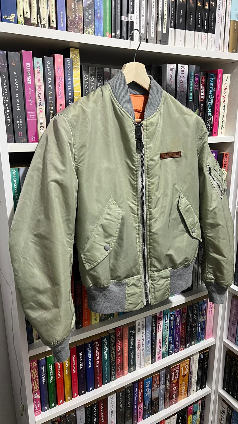 May include: A sage green bomber jacket featuring a ribbed gray collar, cuffs, and hem. The jacket has a full-length front zipper, two side pockets with snap closures, a small zippered utility pocket on the left sleeve, and a rectangular patch.