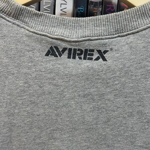 May include: A gray crew neck sweatshirt with the brand name "AVIREX" printed in black across the chest. The sweatshirt is hanging on a wooden hanger. The background includes a shelf with books.