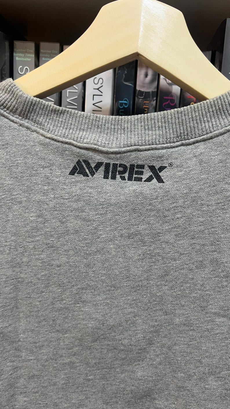 May include: A gray crew neck sweatshirt with the brand name "AVIREX" printed in black across the chest. The sweatshirt is hanging on a wooden hanger. The background includes a shelf with books.